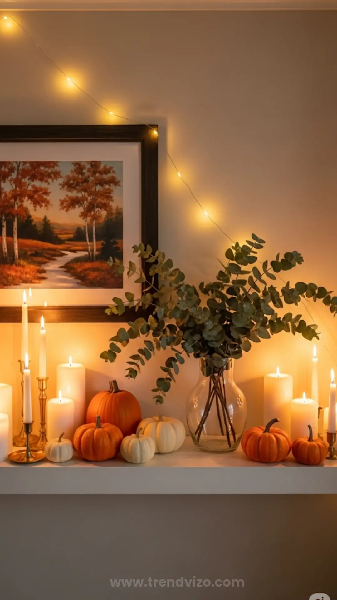 A cozy autumn mantel display featuring candles, small orange and white pumpkins, a vase with green foliage, string lights, and a fall landscape painting.