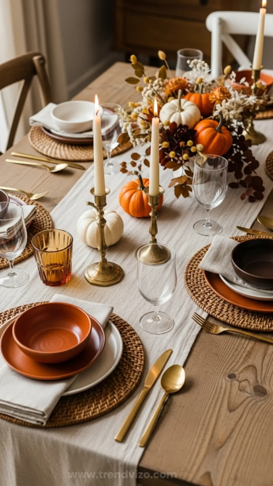 Elegant autumn table setting with white and orange pumpkins, brass candlesticks, and woven placemats. Warm, cozy ambiance with fall-themed decor.