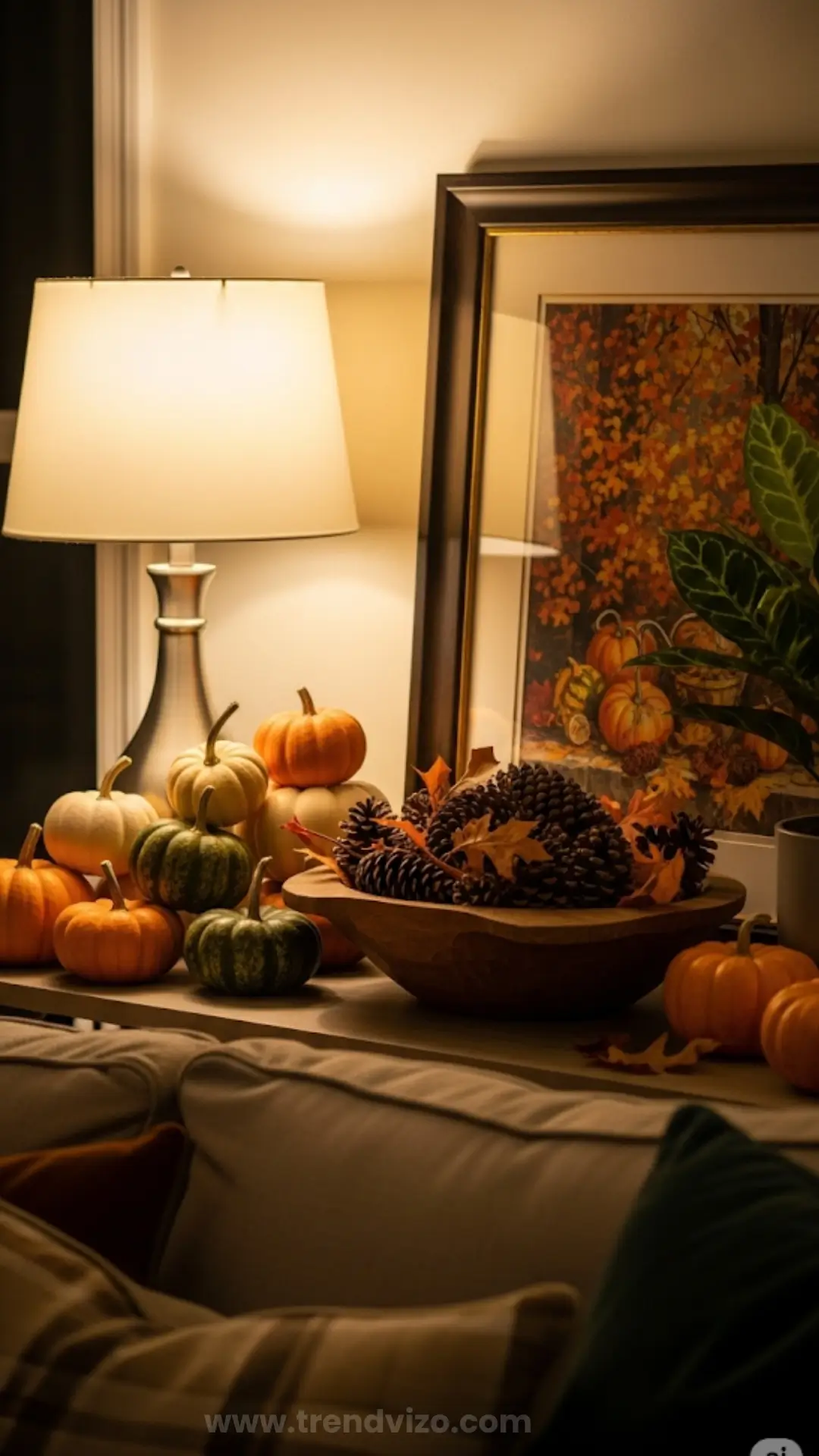 Cozy autumnal scene with small pumpkins and pinecones on a shelf, soft lamp lighting, a fall-themed painting, and lush plant, evoking warmth.